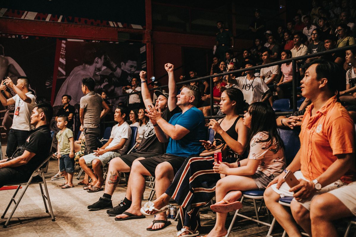 A mixed crowd of tourists and Thai locals in bleacher seating, several mid-cheer, one man fist-pumping.