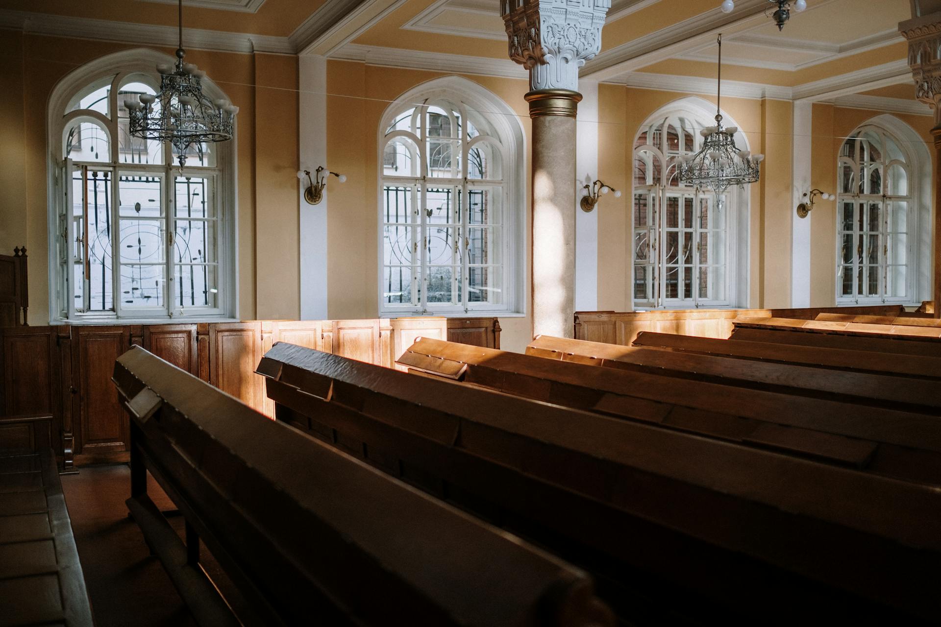 Sunlit church interior with arched windows, wooden pews, and chandeliers.