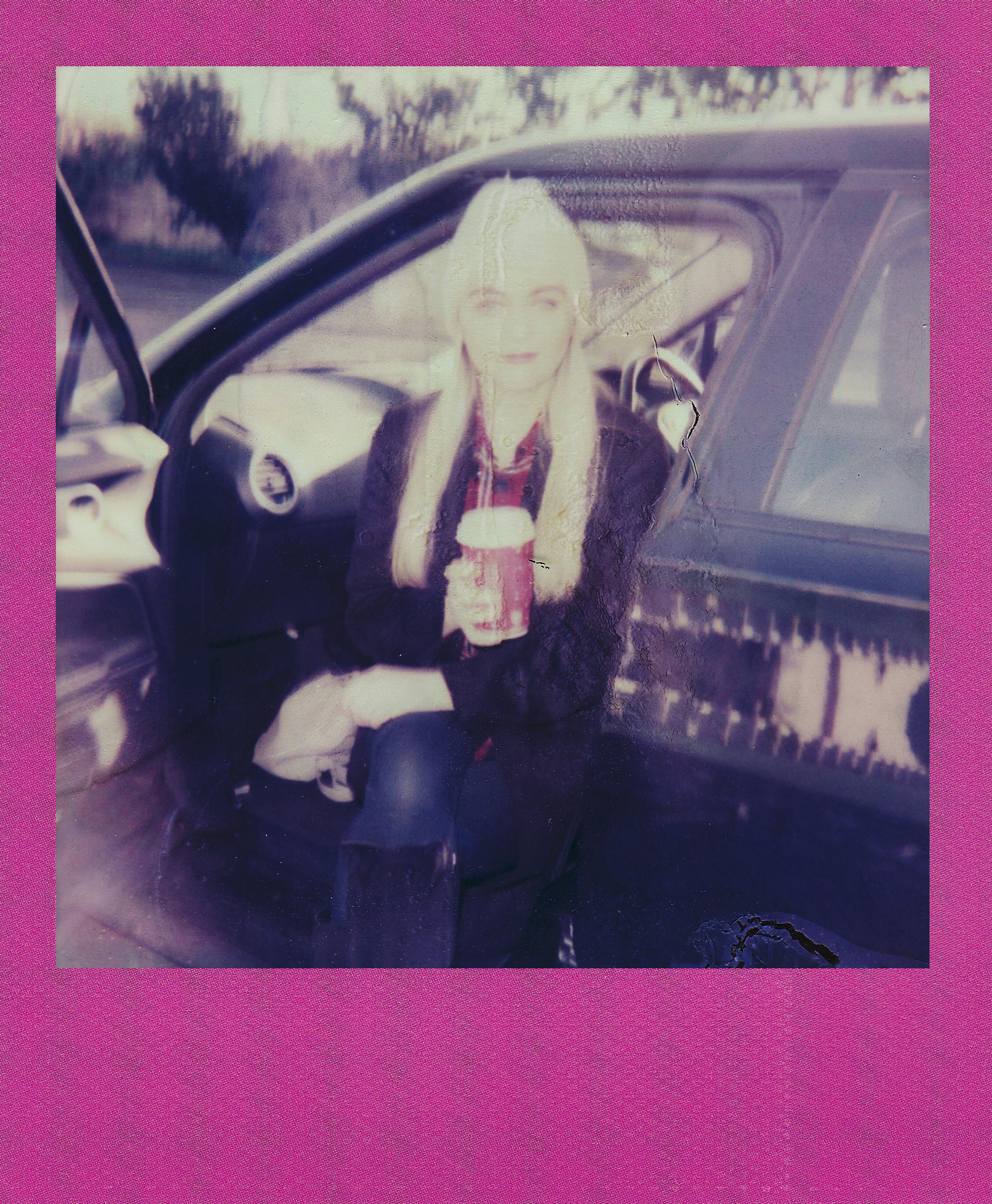 A young woman with long hair sits by a car holding a warm drink, capturing a relaxed moment.