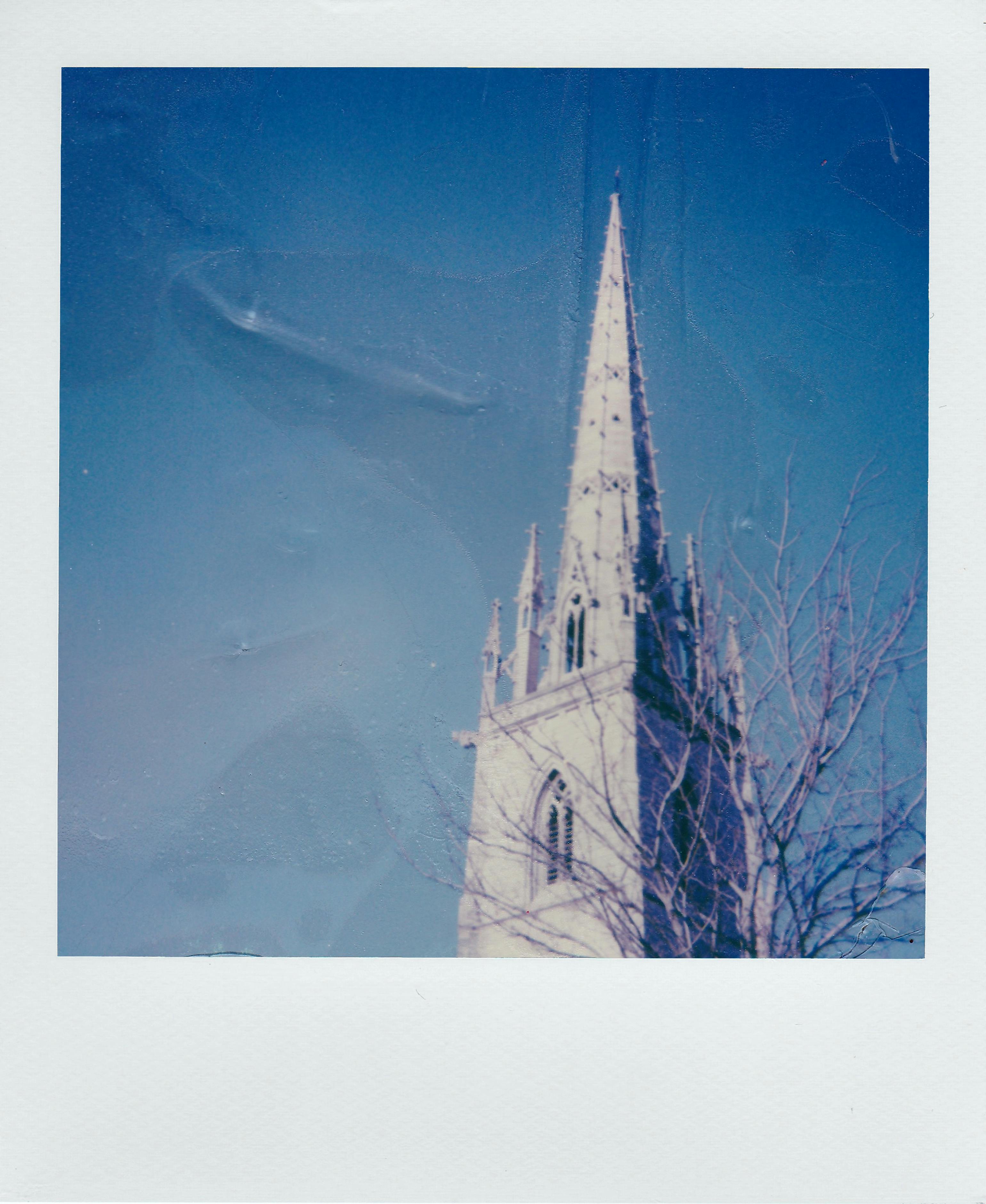 A Gothic church spire stands tall against a clear blue sky, framed by a bare tree.