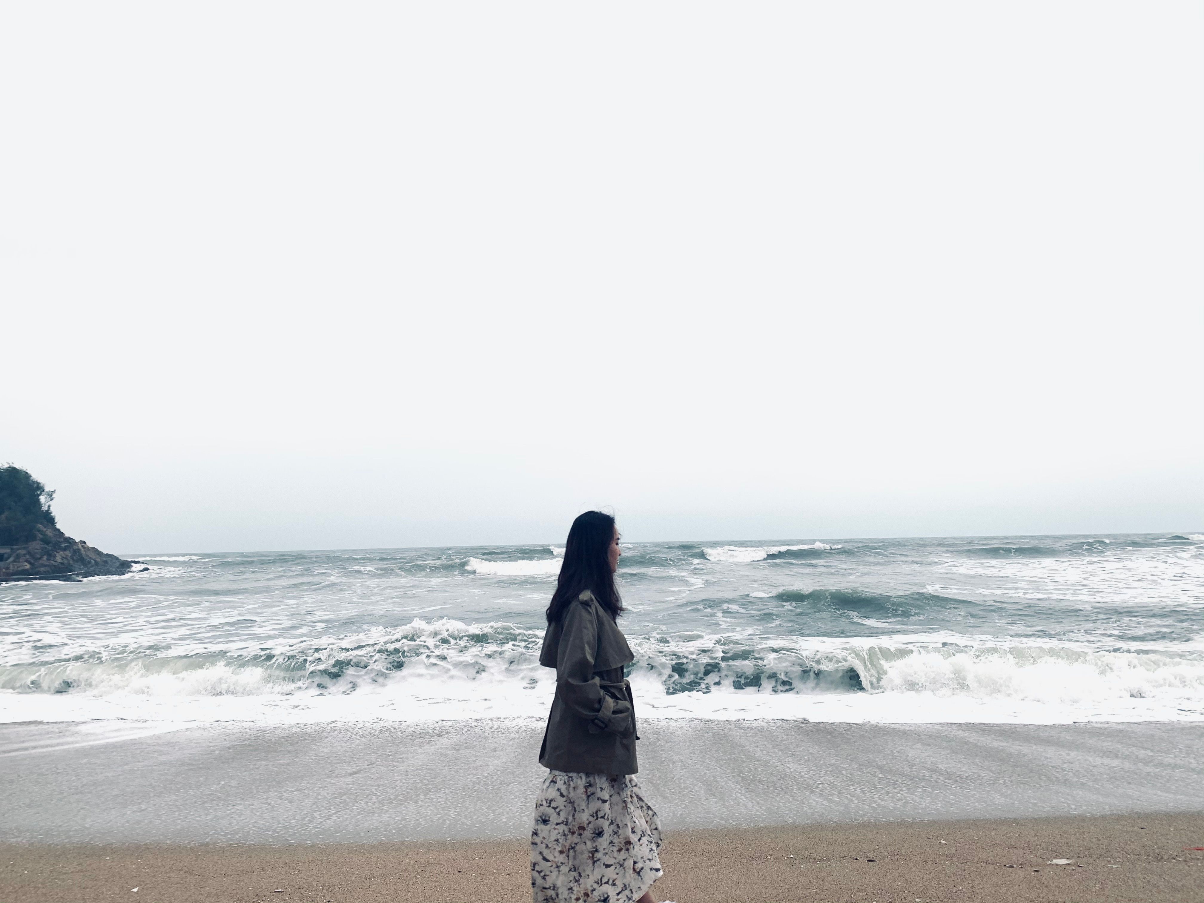 A woman walks on a quiet beach with waves crashing and a cloudy sky overhead.
