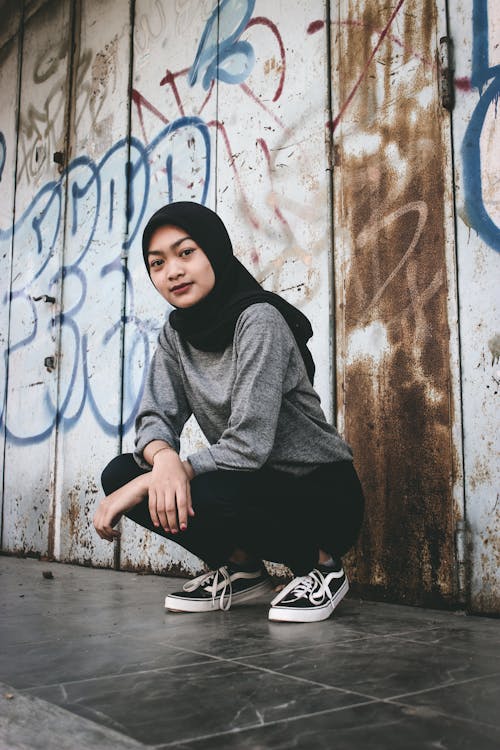 Woman in Black Hijab Squatting Beside a Wall with Graffiti