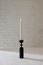 Burning candle on black candlestick placed on table against white brick wall in light room with shadow