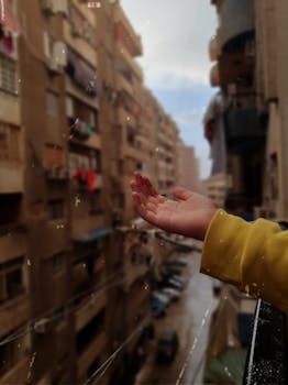 A child's hand reaching out from a balcony during rainfall in a city setting, capturing the moment's tranquility.