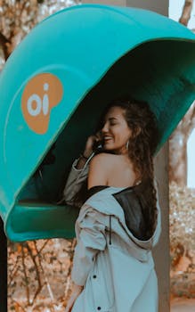 Young woman smiling while making a call from a retro public phone booth outdoors.