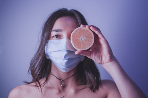Young female in medical mask looking at camera and covering eye with half of orange while standing near wall during pandemic