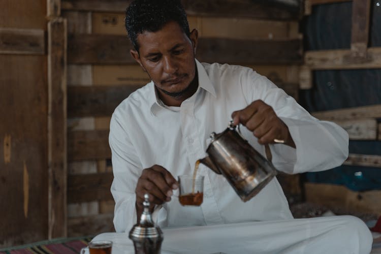 A Man In A White Thobe Pouring Tea From A Kettle