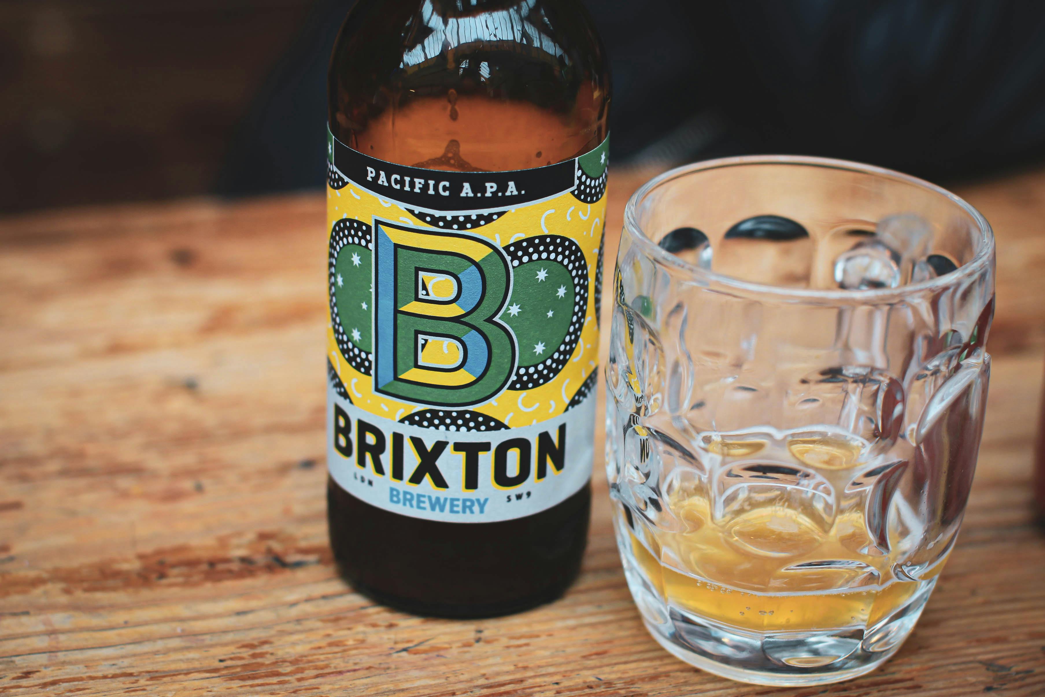 Close-up of Brixton Brewery's Pacific APA beer bottle with a glass on a wooden table.