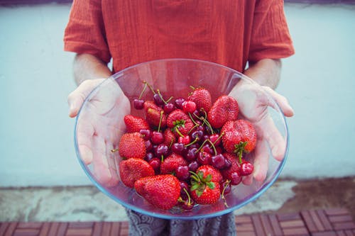 From above crop anonymous person in casual clothes demonstrating glass bowl filled with ripe yummy strawberries and cherries in backyard