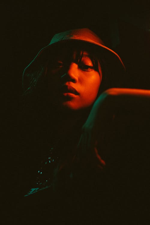 Free Young ethnic female in hat standing in dark place and looking at camera Stock Photo