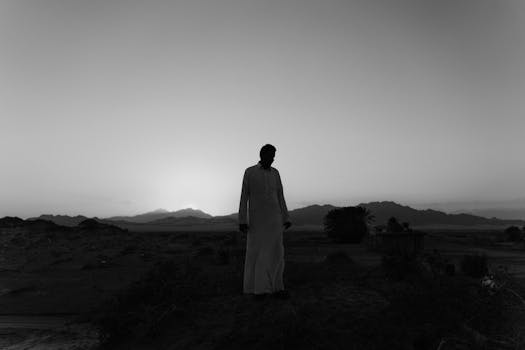 Silhouette of a man in traditional attire against desert mountains at sunrise, in monochrome.