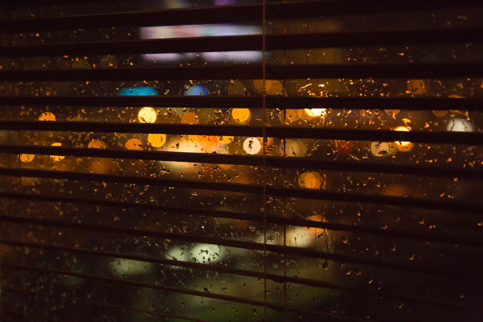 Raindrops on window blinds create a bokeh effect, capturing a moody cityscape at night.