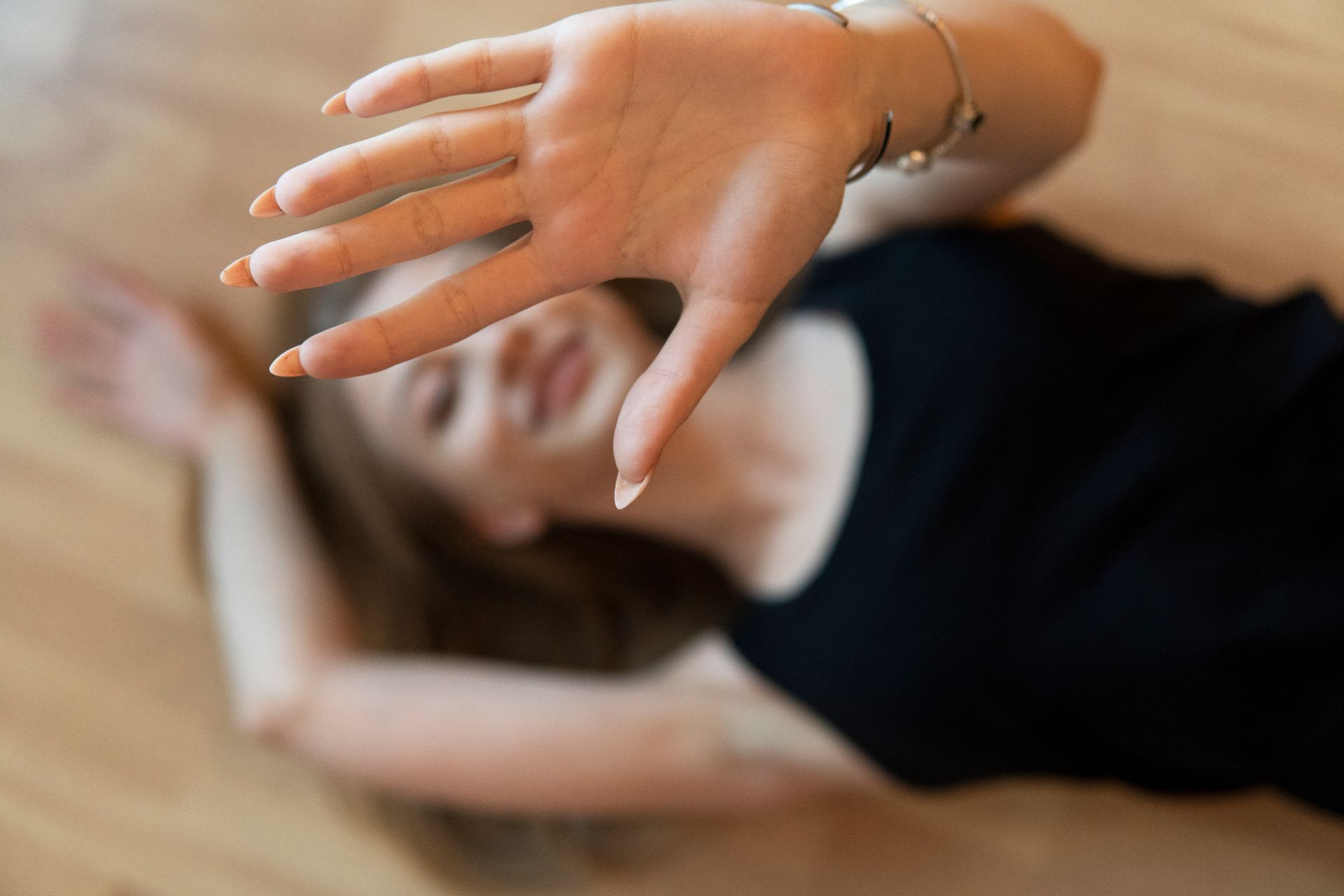 A woman lying on a wooden floor with a hand gesture blocking the frame, smiling naturally.