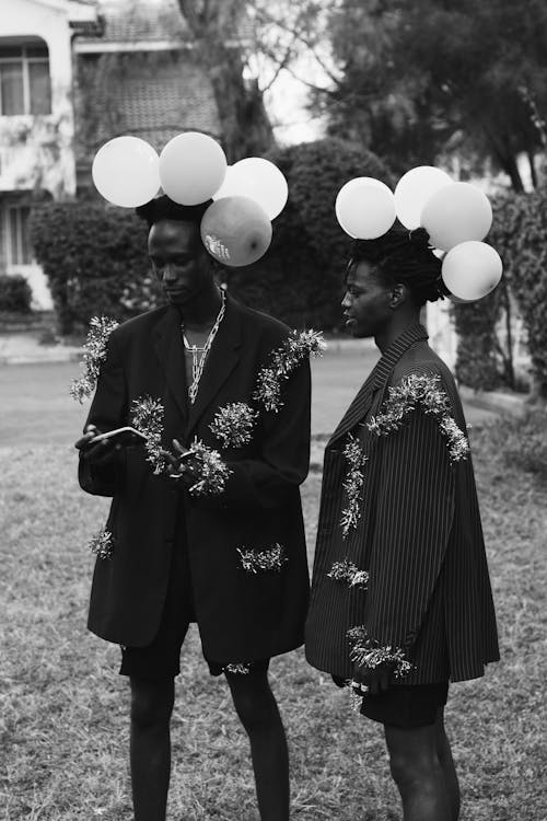 Black and white of African American male friends with balloons on head in suits with tinsel browsing mobile phone while standing on backyard