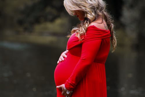 Free Side view of wavy haired female in red dress holding hands on tummy while standing against water Stock Photo