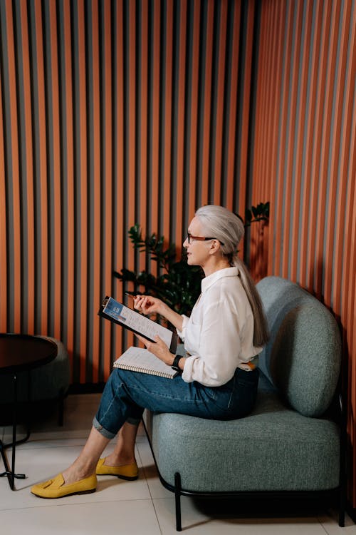 Woman in White Long Sleeve Shirt and Blue Denim Jeans Sitting on Gray Sofa Chair