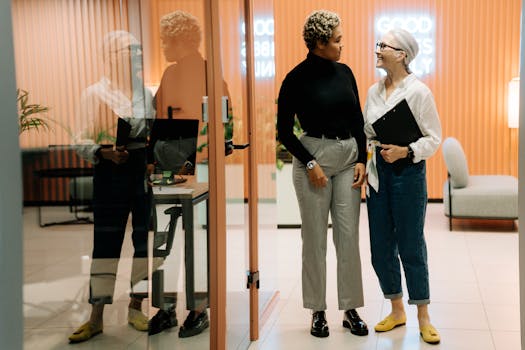 Two professional women discussing in a modern office with a reflective glass wall.