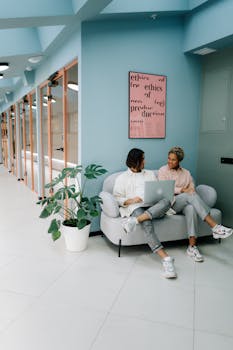 Two colleagues discussing work on a laptop in a modern office corridor.
