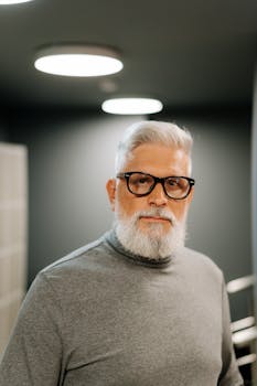 A senior man with a beard and glasses posing indoors, exhibiting modern style and elegance.