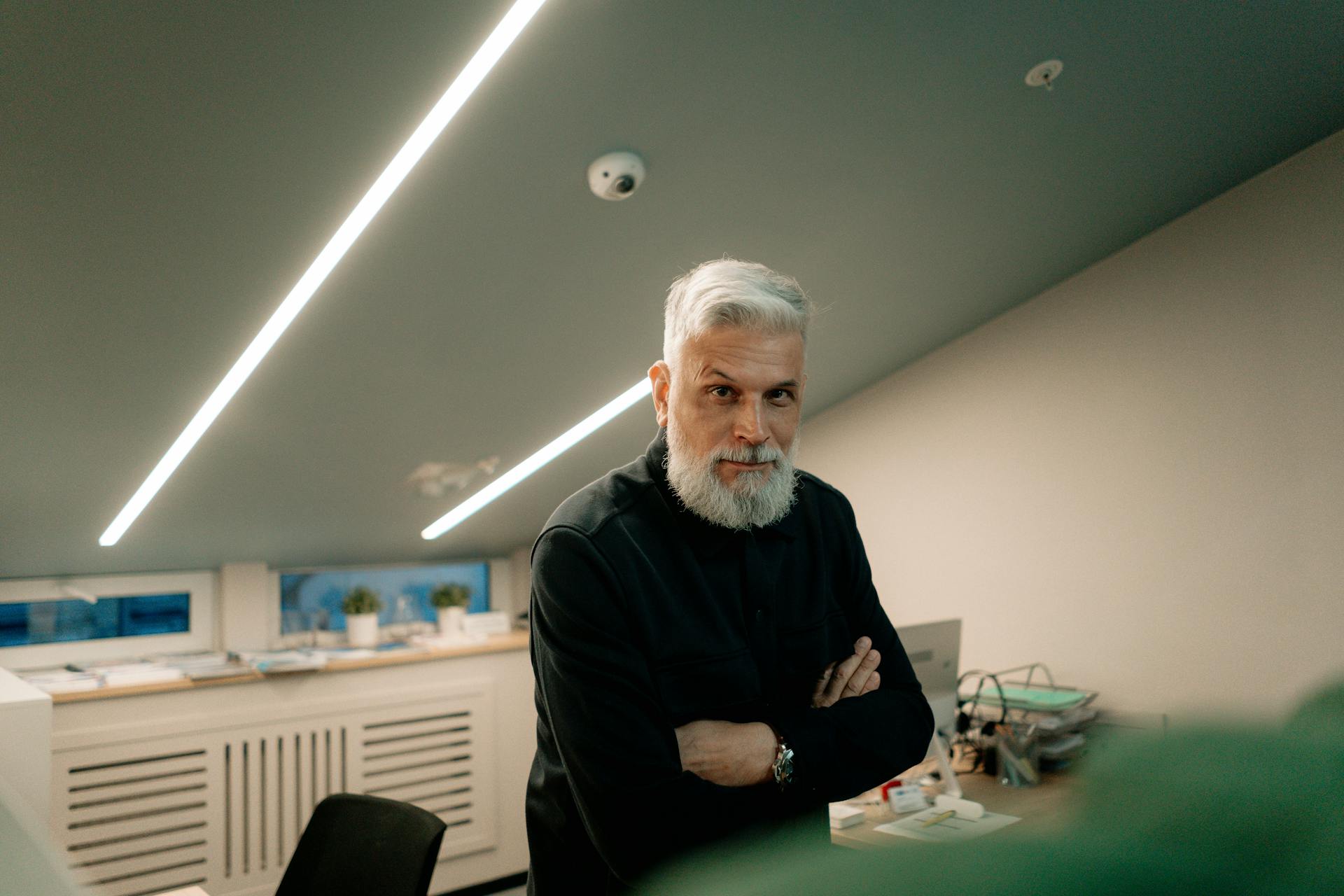 A confident mature man with gray hair and beard, standing in a modern office environment.