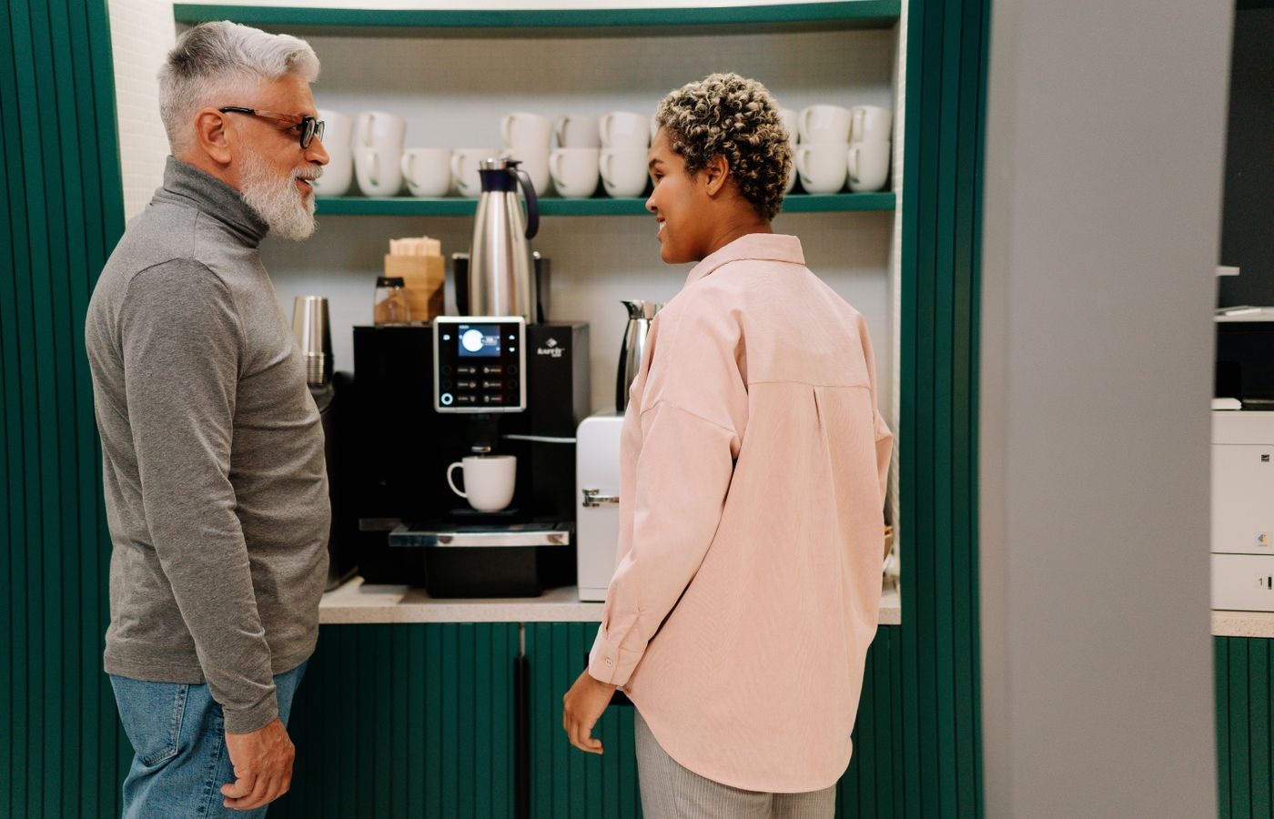 Senior and adult colleagues enjoying a coffee break, chatting by the coffee machine in an office break room.