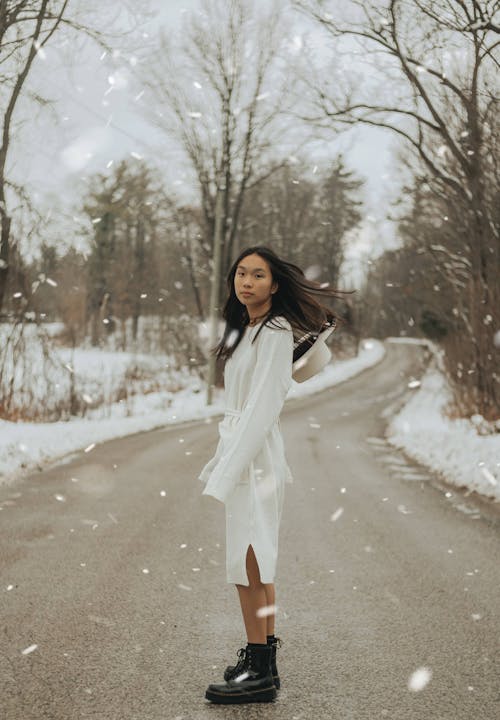 Side view of attractive ethnic female in dress standing on asphalt roadway in snowy weather