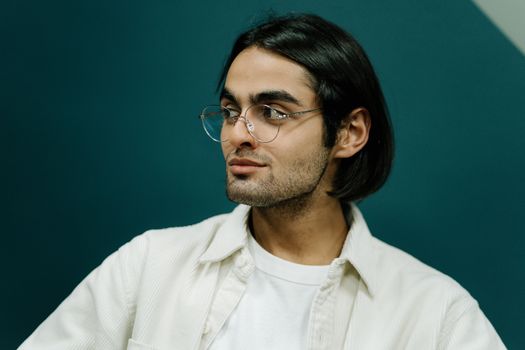 A thoughtful portrait of a stylish man wearing eyeglasses against a teal background.