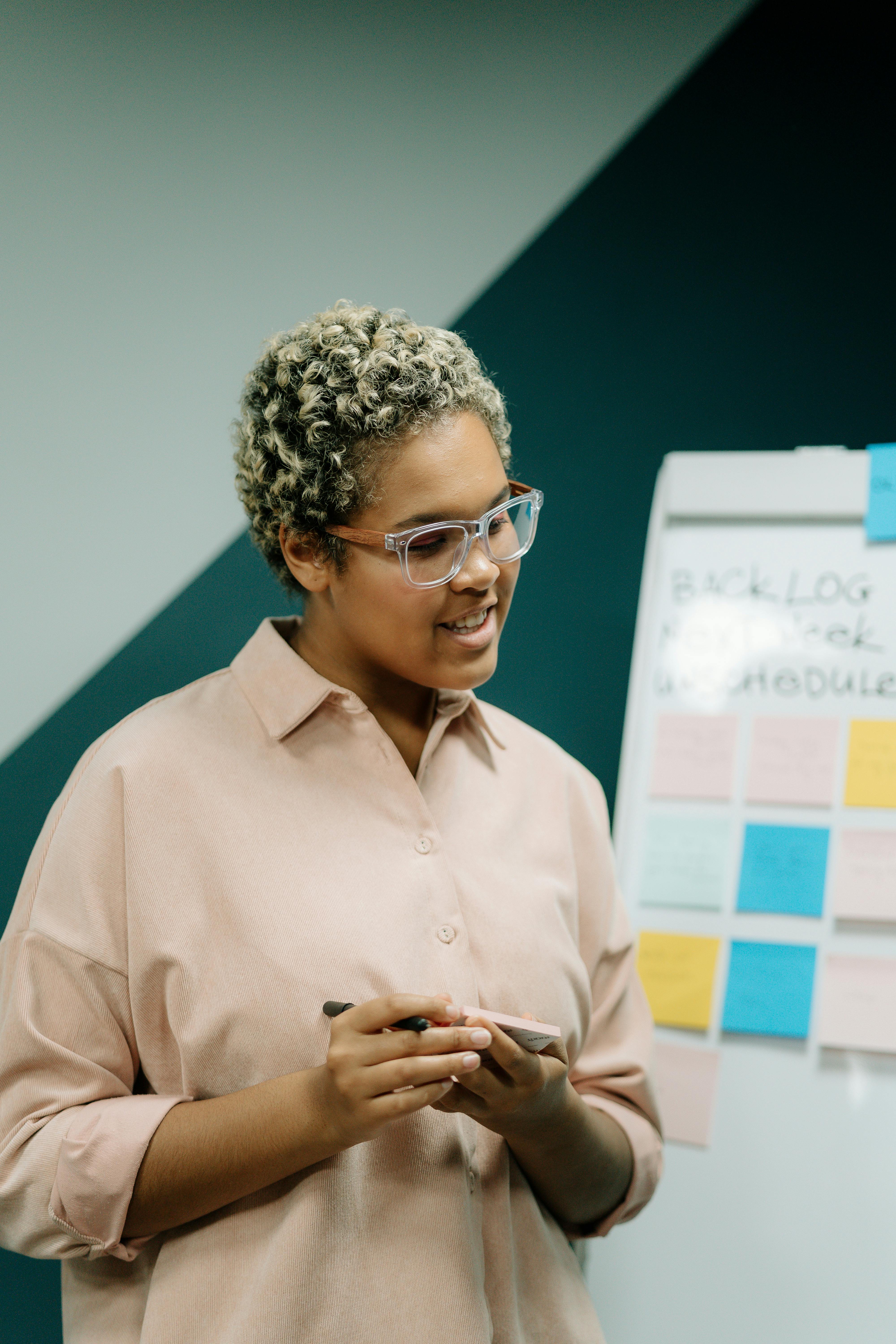 woman in beige shirt wearing eyeglasses and standing beside white board with stickers