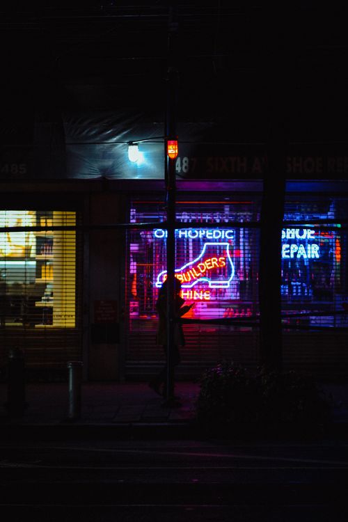 Free Side view full body of unrecognizable woman on sidewalk near buildings with glowing neon signboards and fence near asphalt road at night in city street Stock Photo
