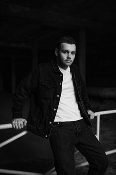 Moody black and white portrait of a man leaning on a railing in a denim jacket.