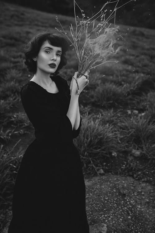 Grayscale Photo of a Woman in Black Dress Holding Dried Grass