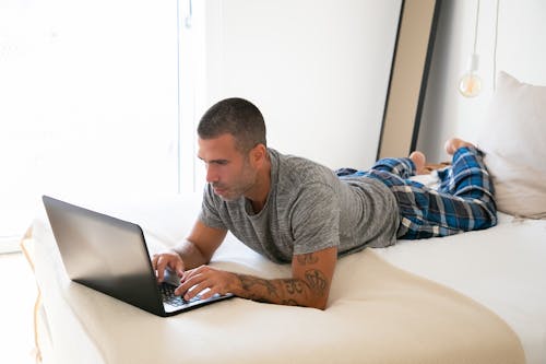 Free Man in Gray Shirt Lying on Bed While Using Laptop Stock Photo