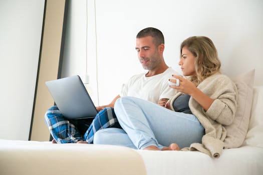A couple sitting on a bed using a laptop, representing work-from-home lifestyle.