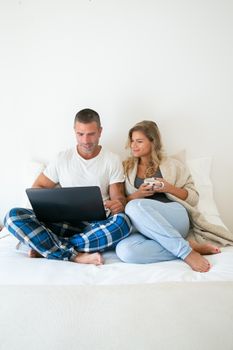 A couple enjoying remote work from home, sitting on bed with a laptop and a mug in comfortable attire.