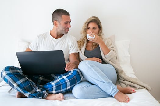 A couple enjoying coffee and using a laptop while relaxing in bed, portraying a cozy work-from-home scenario.