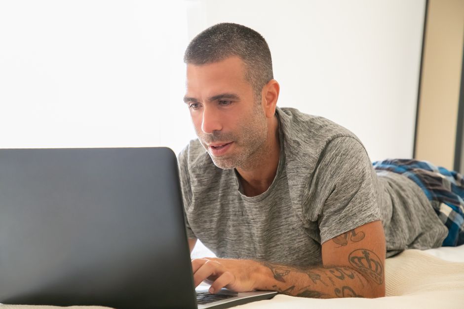 Nectar vs Tuft and Needle: Memory Foam Showdown for Budget Buyers An adult man working remotely on a laptop while lying on his bed at home, showcasing a relaxed work environment.