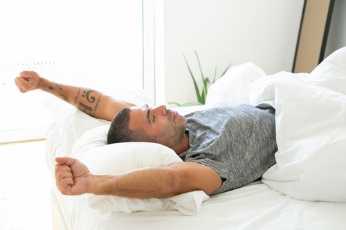 Free Adult man stretching in bed, looking relaxed and refreshed in the morning light. Stock Photo