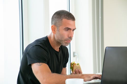 Free Man Holding a Sandwich While Using a Laptop Stock Photo