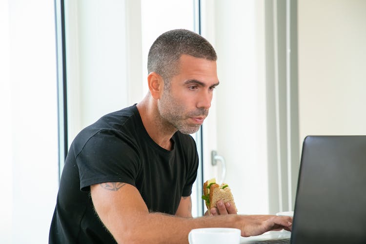 Man Holding A Sandwich While Using A Laptop