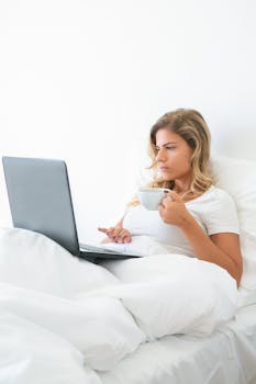 A woman sitting comfortably in bed, working on a laptop with coffee.