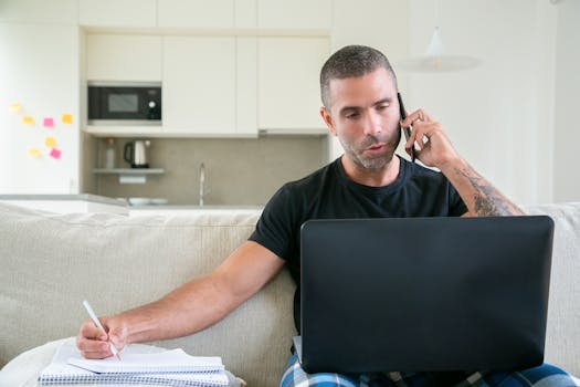 Man working from home using a laptop and smartphone while taking notes in a modern kitchen setting.