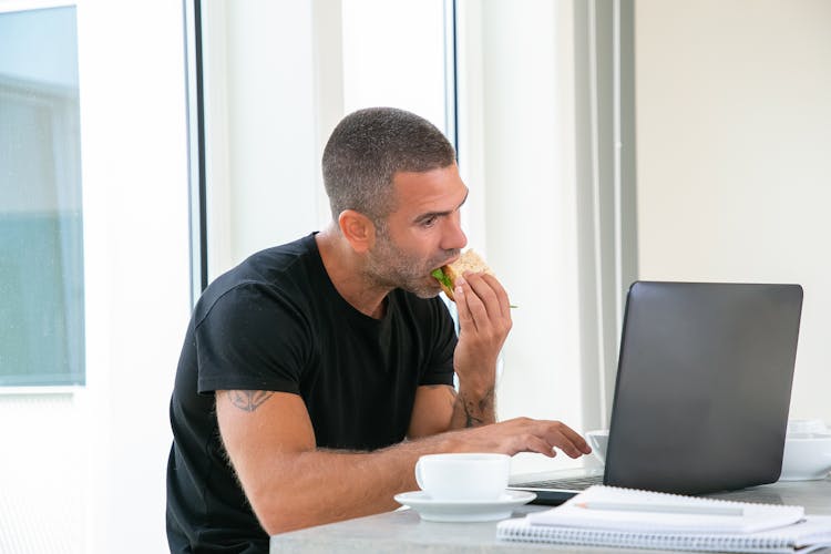 Man In Black Shirt Eating Sandwich While Using His Laptop