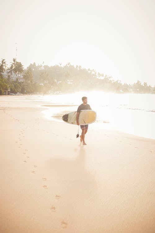 Free Man Carrying Surfboard While Walking on Shore Stock Photo
