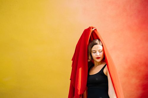 Free A contemplative woman draped in a red cloth stands against a gradient yellow and orange backdrop. Stock Photo