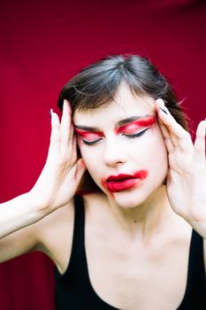 Close-up of a woman with eyes closed and smeared red makeup, holding her temples against a vivid red background.