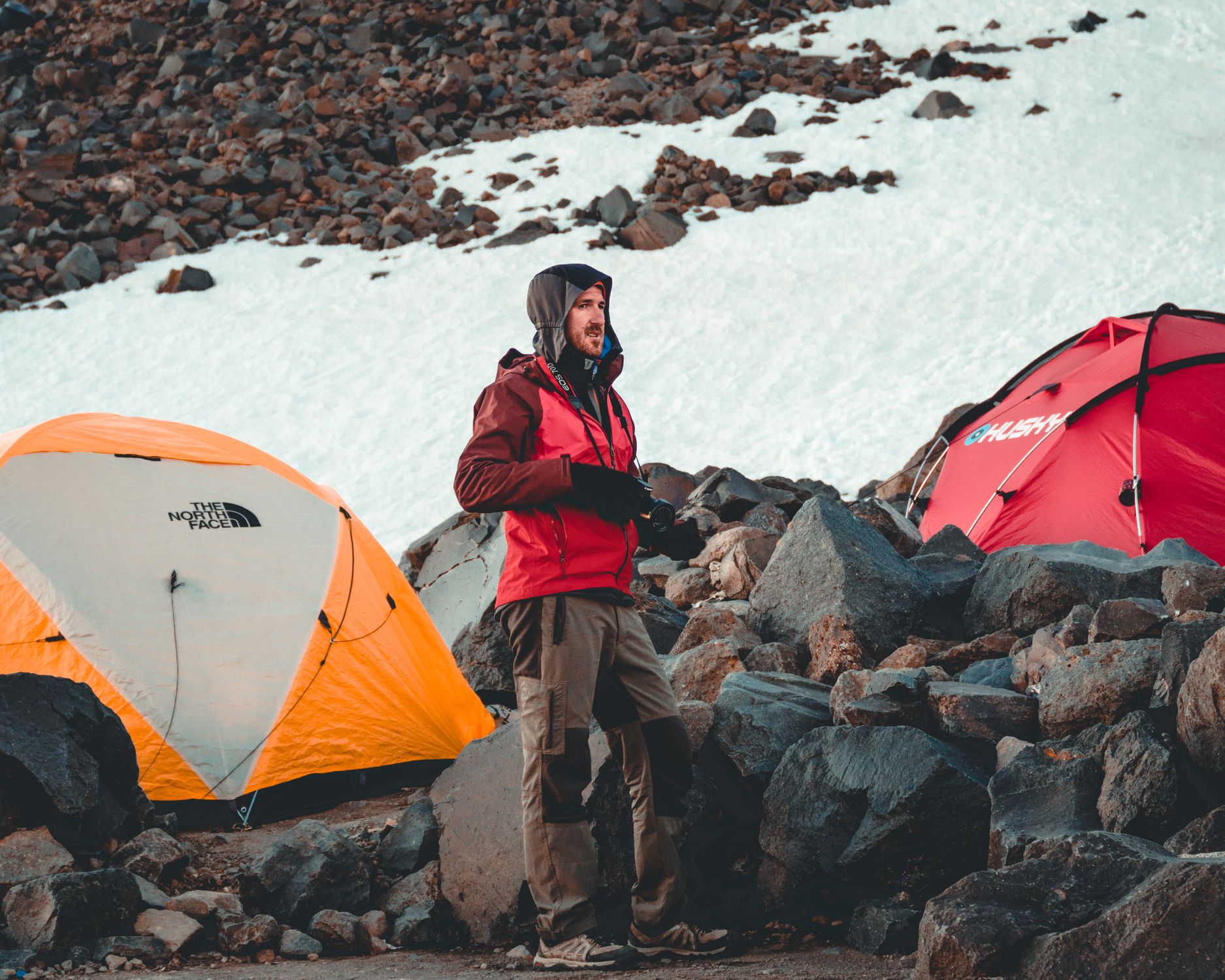Man camping in snow-covered mountains with tents and rocky terrain.