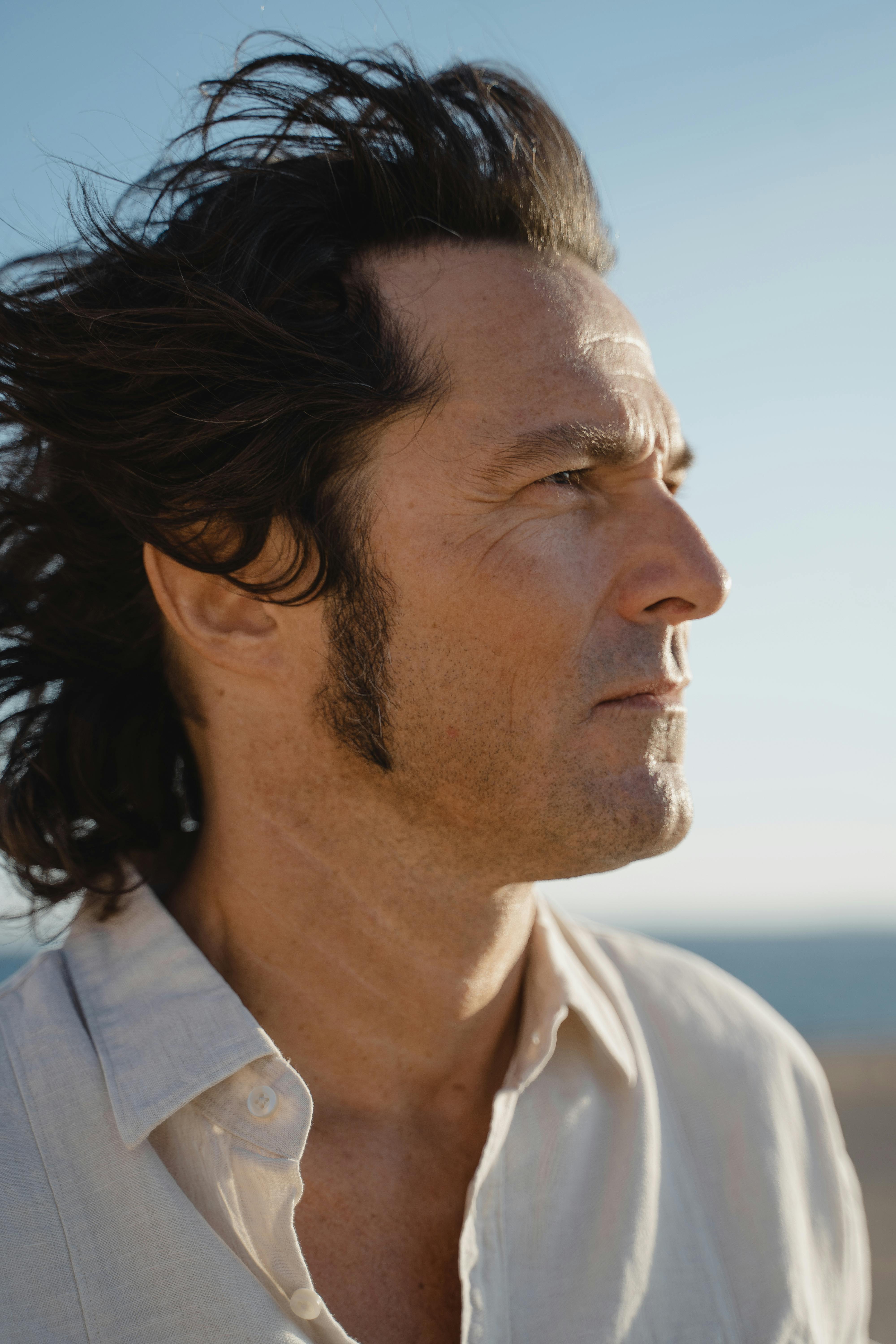 A thoughtful side profile portrait of a man with windswept hair, outdoors under a clear sky.
