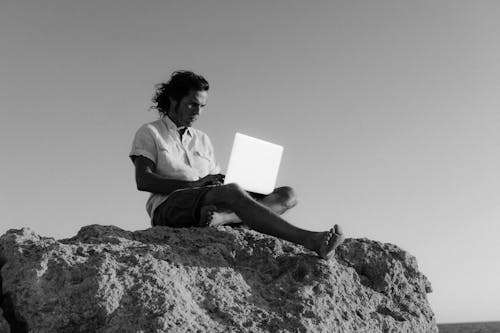 Man in White Dress Shirt Sitting on Rock Using a Laptop