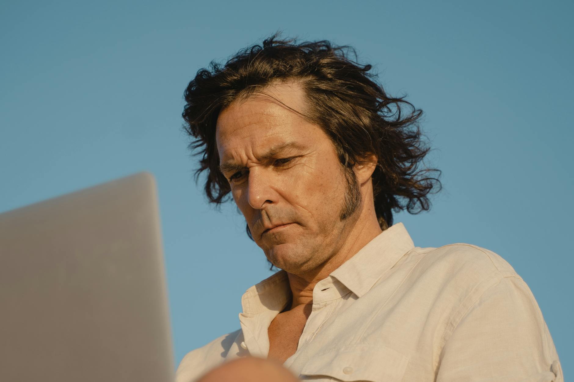 A man with long hair intensely focusing on his laptop under a clear blue sky.