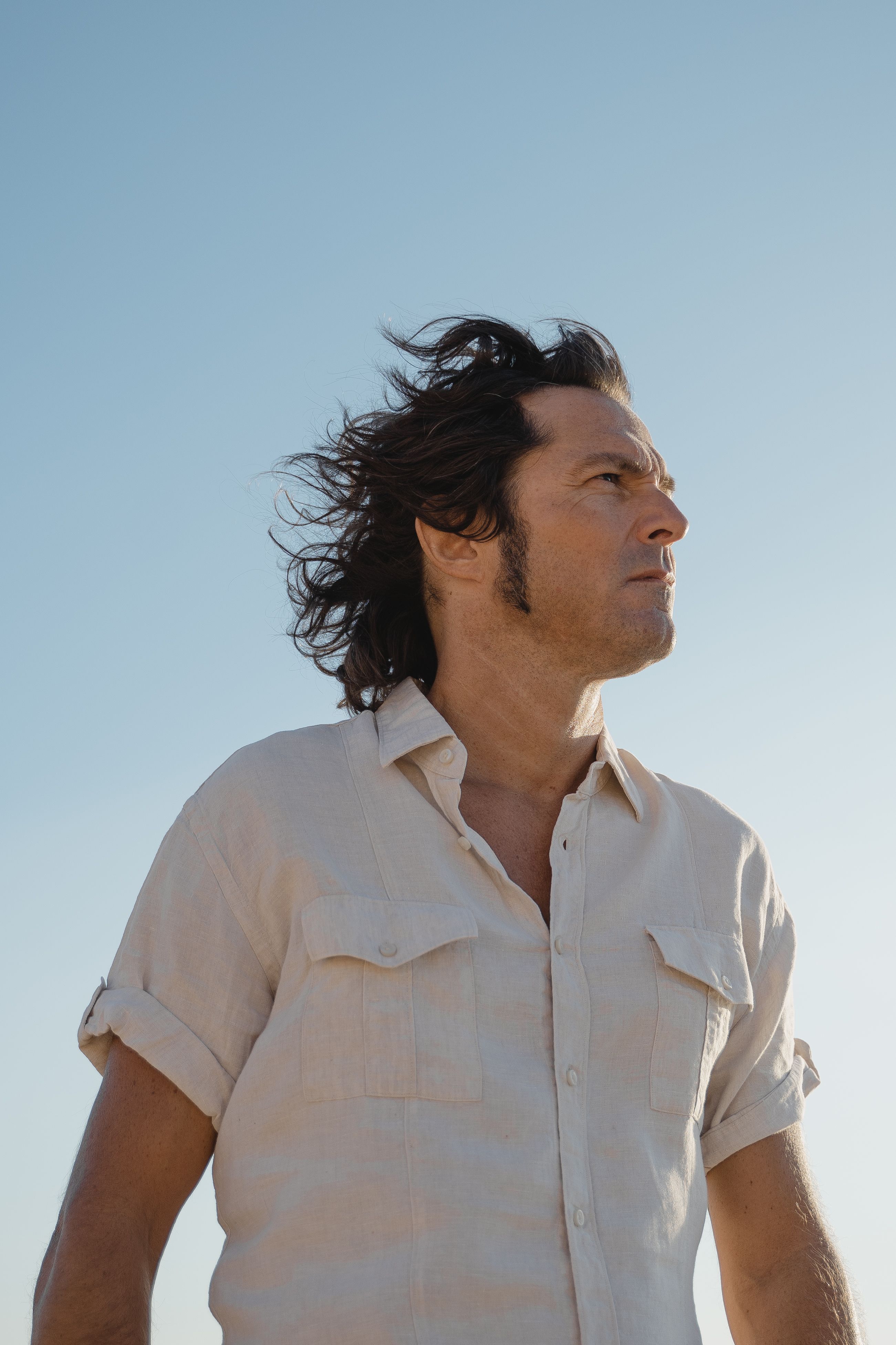 Side view of a man with windswept hair in a relaxed pose outdoors against a clear blue sky.